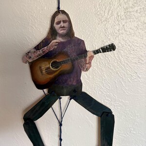 May include: A wooden, articulated figure of a person playing an acoustic guitar. The figure is painted with a purple shirt, dark pants, and white-soled shoes. The guitar is brown with a detailed sound hole. The figure is suspended by a blue string.