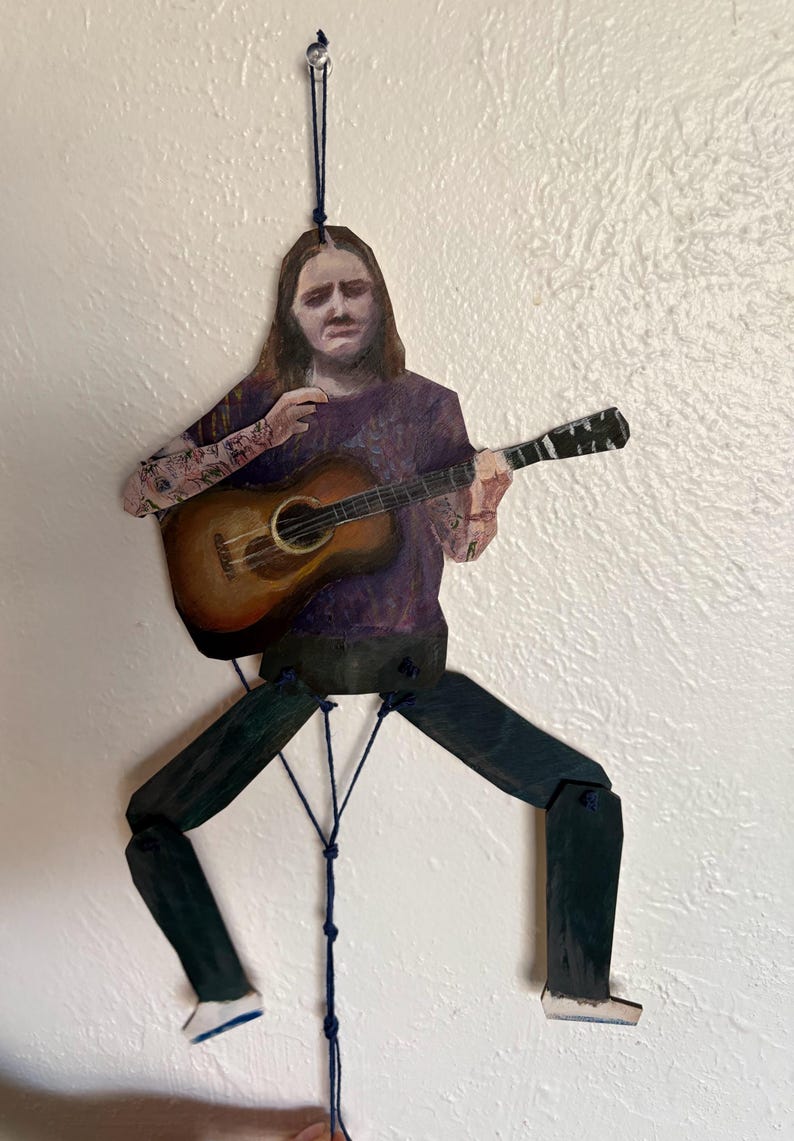 May include: A wooden, articulated figure of a person playing an acoustic guitar. The figure is painted with a purple shirt, dark pants, and white-soled shoes. The guitar is brown with a detailed sound hole. The figure is suspended by a blue string.