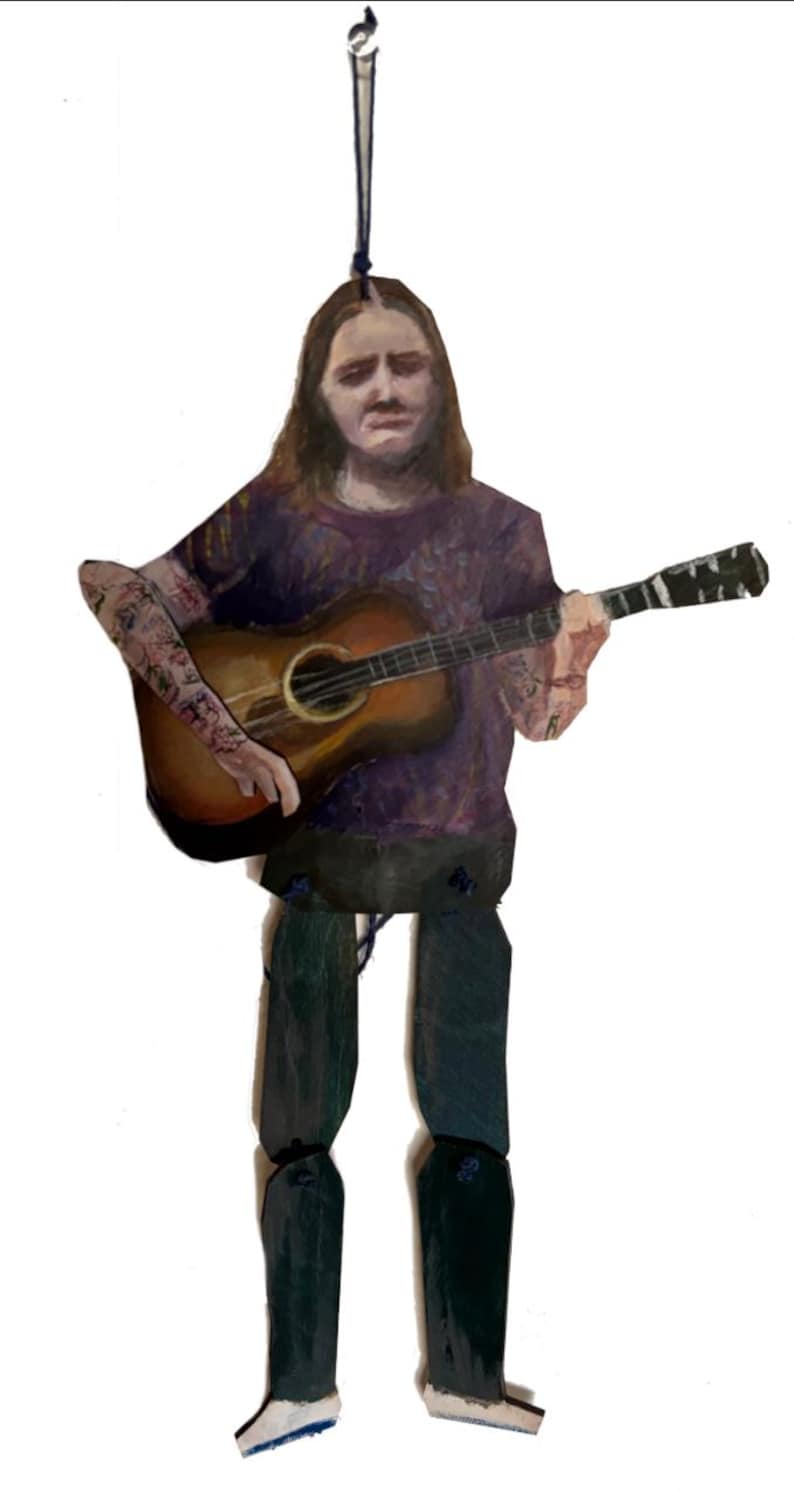 May include: Wooden ornament depicting a person playing an acoustic guitar. The figure wears a purple shirt and dark pants. The guitar is brown with a black fretboard. The ornament is suspended by a black string.