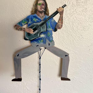 May include: Wooden articulated figure of a person playing a guitar. The figure has long blonde hair, sunglasses, and tattoos. It wears a blue Hawaiian shirt and grey trousers. The figure is suspended by a dark blue string.