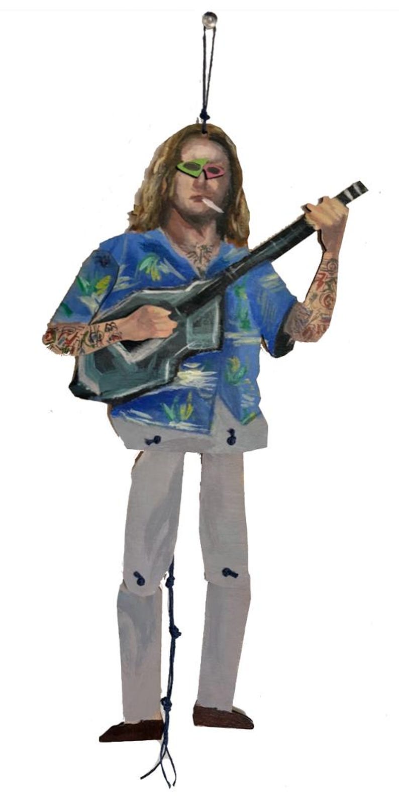 May include: Wooden ornament depicting a person with long hair, sunglasses, and tattoos, playing a guitar. The person wears a blue Hawaiian shirt and white trousers. The ornament hangs from a black string.