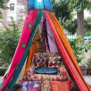 Może przedstawiać: Kolorowa strefa wypoczynkowa na świeżym powietrzu w stylu boho. Fotel w wzory zdobią poduszki w różnych kolorach i wzorach. Drapowany baldachim z żywych tkanin, w tym różu, pomarańczy i błękitu, tworzy przytulną, zacienioną przestrzeń.