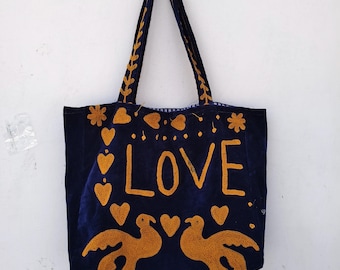 May include: A navy blue tote bag with gold embroidered details. The word "LOVE" is prominently displayed, along with hearts, flowers, and two birds. The handles and top edge feature decorative stitching.