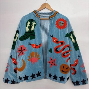 May include: A light blue velvet jacket with colorful embroidered designs. The jacket features a green cowboy boot, a red snake, a smiling sun, and other whimsical shapes. The interior lining has a striped pattern.