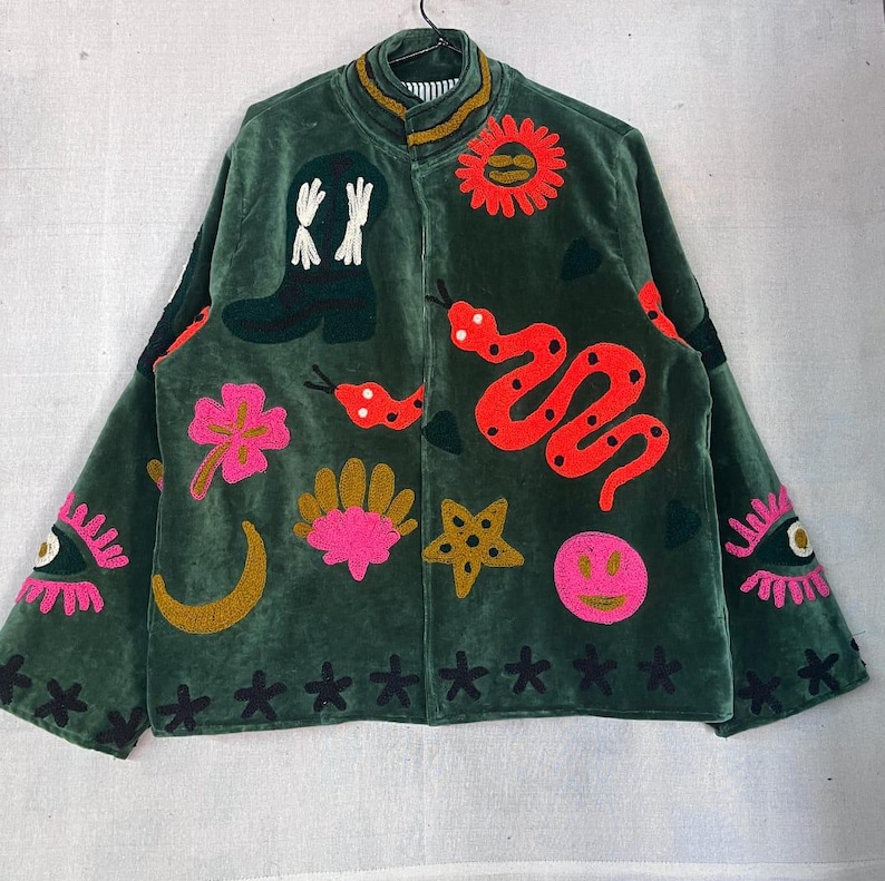 May include: A dark green velvet jacket featuring colorful embroidered motifs including a cowboy boot, a red snake, a sun, a pink flower, a gold crescent moon, a shell, a star, a smiley face, and eyes on the sleeves, with a high collar.