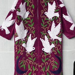 May include: A long, magenta velvet coat with white dove and green vine embroidery. The coat features a high collar and a striped lining. The design includes crescent moons at the bottom. A unique, artistic garment.
