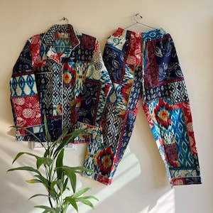 Peut inclure: Un ensemble de pyjama deux pièces avec un motif patchwork. L'ensemble comprend une chemise boutonnée à manches longues et un pantalon assorti. Le tissu présente un mélange de motifs rouges, bleus et crème. Le pyjama est suspendu à un cintre.