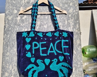 May include: A navy blue tote bag with teal embroidered details. The word "PEACE" is embroidered in large teal letters. The bag features teal hearts, flowers, and two birds. The handles are teal with a leaf design.