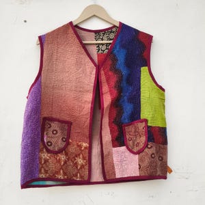 May include: A patchwork vest in a variety of colors, including purple, pink, red, blue, and green. The vest has two pockets and a V-neck. The edges are trimmed in a dark red fabric. The vest is hanging on a wooden hanger.