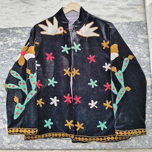 May include: Black velvet jacket with colorful embroidered floral and cactus designs. Features gold trim and a striped lining. Embellished with white birds and star-shaped accents. A unique, eye-catching piece.
