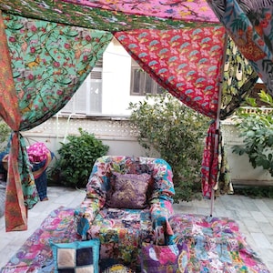 Puede incluir: Una colorida zona de estar al aire libre con un sillón patchwork, cojines y una manta estampada. El dosel superior está hecho de paneles de tela vibrantes con estampados florales y de animales. La escena está ambientada en un patio con vegetación.