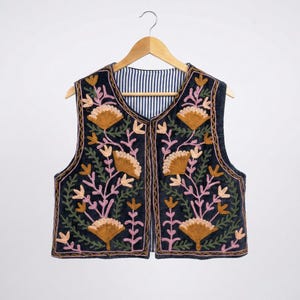 May include: A sleeveless, dark navy vest with an embroidered floral design. The vest features a detailed pattern of gold, pink, and green flowers and leaves. The interior lining has a blue and white striped pattern. The vest is hanging on a wooden hanger.