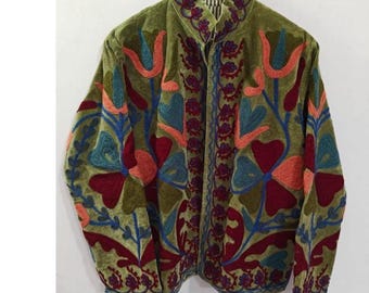 May include: Olive green velvet jacket with colorful floral embroidery. The jacket features a high collar and is adorned with red, orange, blue, and purple floral patterns. The jacket is hanging on a wooden hanger.