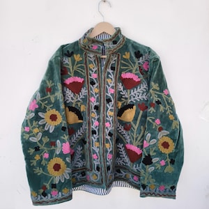 May include: A teal velvet jacket with floral embroidery in pink, yellow, and black. The jacket features a high collar and a striped lining. The embroidery includes various flower and leaf designs, creating a detailed and colorful pattern.