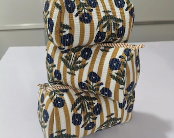 Pode incluir: Três nécessaires retangulares empilhadas. Cada nécessaire apresenta um padrão de listras brancas e amarelo mostarda com detalhes florais azuis. As nécessaires têm uma borda marrom e uma pequena alça lateral.
