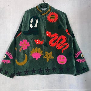 May include: A dark green velvet jacket featuring colorful embroidered motifs including a cowboy boot, a red snake, a sun, a pink flower, a gold crescent moon, a shell, a star, a smiley face, and eyes on the sleeves, with a high collar.