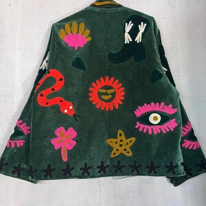 May include: A dark green velvet jacket featuring colorful embroidered motifs including a red snake, a black cowboy boot, a bright orange sun, a pink eye, a yellow starfish, and a pink flower. The hem is decorated with a row of black stitched crosses.