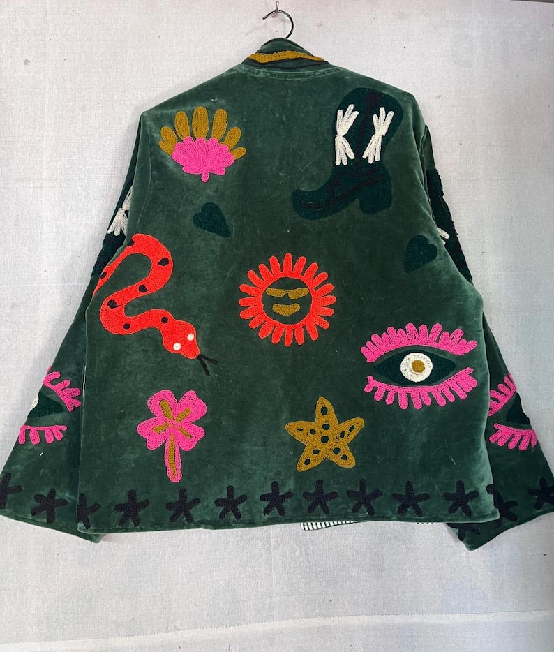 May include: A dark green velvet jacket featuring colorful embroidered motifs including a red snake, a black cowboy boot, a bright orange sun, a pink eye, a yellow starfish, and a pink flower. The hem is decorated with a row of black stitched crosses.