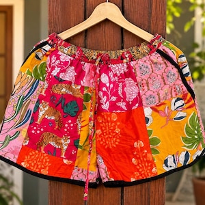 May include: Patchwork shorts featuring a vibrant mix of fabrics with tiger illustrations, floral patterns, and botanical motifs in shades of pink, orange, yellow, and green. The shorts have an elastic waistband with a drawstring and black trim.