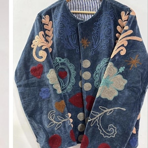 May include: A blue velvet jacket with intricate embroidery in shades of blue, peach, and brown. The jacket features floral and heart-shaped appliqués, and a striped lining. It is displayed on a wooden hanger.