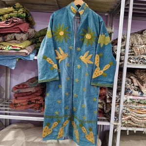 Peut inclure: Long manteau en velours bleu sarcelle à devant ouvert, orné de broderies dorées représentant des soleils, des oiseaux et des motifs floraux. Le manteau a un col mandarin et est doublé d'un tissu rayé.