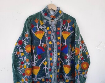 May include: A teal velvet jacket with colorful floral embroidery. The jacket features a mandarin collar and a striped lining. The embroidery includes yellow, orange, red, and blue flowers with white and purple accents. The jacket is hanging on a wooden hanger.