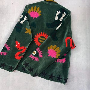 May include: A dark green velvet jacket featuring colorful embroidered motifs including a bright red snake, a yellow sun, pink eyes with long lashes, gold fan shapes, and black cross-stitch patterns scattered across the front and sleeves.