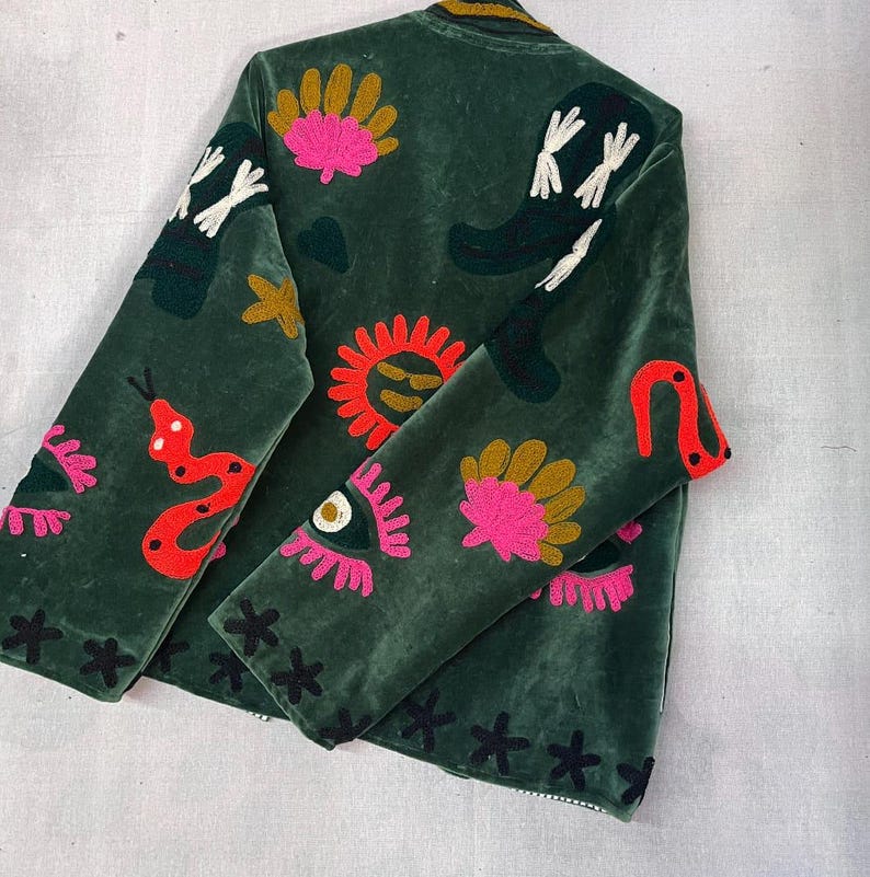 May include: A dark green velvet jacket featuring colorful embroidered motifs including a bright red snake, a yellow sun, pink eyes with long lashes, gold fan shapes, and black cross-stitch patterns scattered across the front and sleeves.