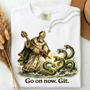 May include: White t-shirt featuring a detailed illustration of a saint with a staff, warding off green snakes. The text "Go on now. Git." is printed at the bottom. A silver necklace with a star pendant is also visible.