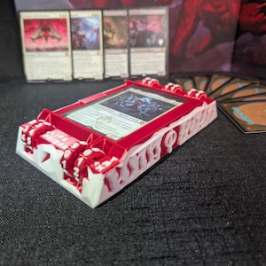 May include: A red and white card holder with a card inside, and a set of cards in the background. The card holder has a red frame and white accents, with a life counter on the side. The card inside has a blue and white image.