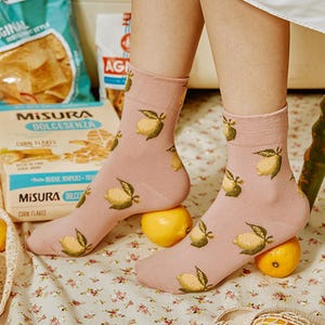 Könnte beinhalten: Hellrosa Socken mit Zitronenmuster. Die Socken zeigen Illustrationen von gelben Zitronen mit grünen Blättern. Die Socken werden mit einem weißen Kleidungsstück getragen und liegen auf Zitronen.