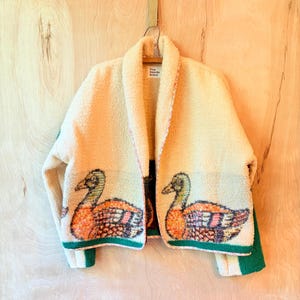 May include: A cream-colored, textured fleece jacket featuring a shawl collar and a repeating graphic of a mallard duck on the lower front panels. The jacket has green trim along the hem and cuffs, with a label reading Fine Needle Brand.