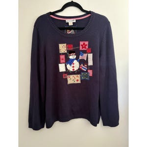 May include: Navy blue long-sleeve sweater with a crew neck. The sweater features a patchwork design with a snowman, flags, and stars. The snowman has a blue scarf and a black top hat. The sweater is hanging on a black hanger.