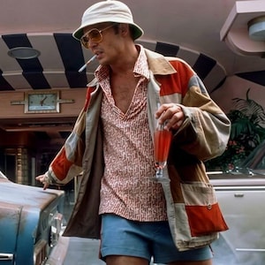 May include: A person wearing a white bucket hat, yellow-tinted sunglasses, a patterned orange and white short-sleeved shirt, and a multi-colored patchwork jacket with red, tan, and grey panels, paired with blue shorts while holding a red drink.