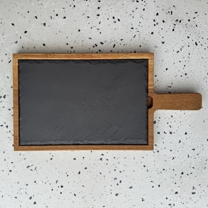 Könnte beinhalten: Rechteckiges Servierbrett mit einer dunkelgrauen Schieferoberfläche und einem Rahmen und Griff aus Naturholz. Geeignet zum Servieren von Vorspeisen oder Käse. Das Holz hat eine warme, hellbraune Farbe.