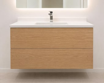 Puede incluir: Un mueble de baño flotante con acabado de madera clara de roble. El mueble tiene dos cajones y una encimera blanca con un lavabo integrado y un grifo cromado. Un espejo retroiluminado está encima del mueble.
