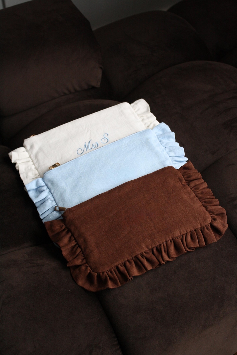 K&ouml;nnte beinhalten: Drei rechteckige Kosmetiktaschen mit R&uuml;schenkanten in Wei&szlig;, Hellblau und Braun. Die wei&szlig;e Tasche hat den gestickten Text "Mrs S" in Hellblau. Die Taschen sind auf einer dunkelbraunen Oberfl&auml;che angeordnet.