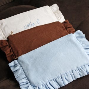 K&ouml;nnte beinhalten: Drei rechteckige Kosmetiktaschen in Wei&szlig;, Braun und Hellblau, jede mit einem ger&uuml;schten Rand. Die wei&szlig;e Tasche hat die blaue Stickerei "Mrs S". Die Taschen sind auf einer braunen Oberfl&auml;che angeordnet.
