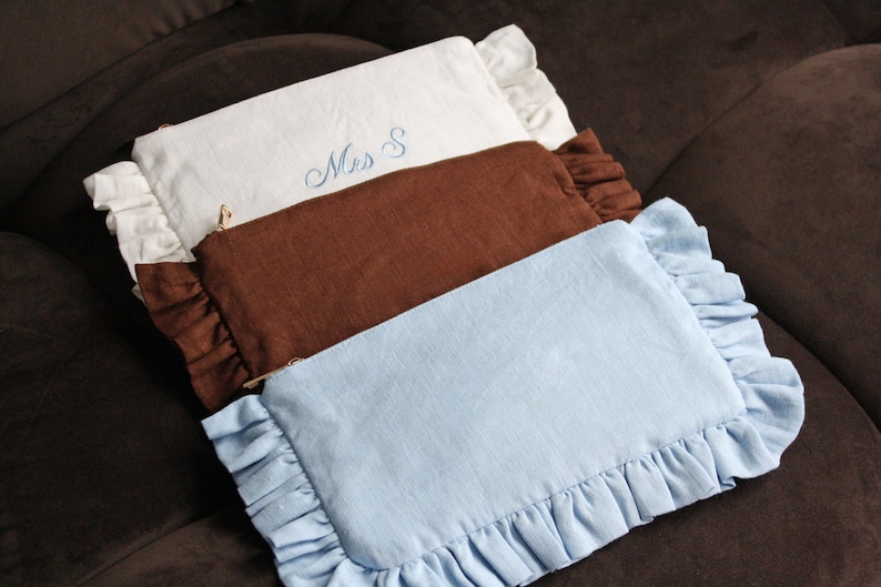 K&ouml;nnte beinhalten: Drei rechteckige Kosmetiktaschen in Wei&szlig;, Braun und Hellblau, jede mit einem ger&uuml;schten Rand. Die wei&szlig;e Tasche hat die blaue Stickerei "Mrs S". Die Taschen sind auf einer braunen Oberfl&auml;che angeordnet.