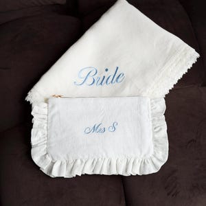 K&ouml;nnte beinhalten: Ein wei&szlig;es Braut-Set aus Leinen. Es besteht aus einem Kissenbezug mit dem blauen Schriftzug "Bride" und einer passenden Clutch mit der blauen Stickerei "Mrs S". Die Clutch hat einen R&uuml;schenrand.
