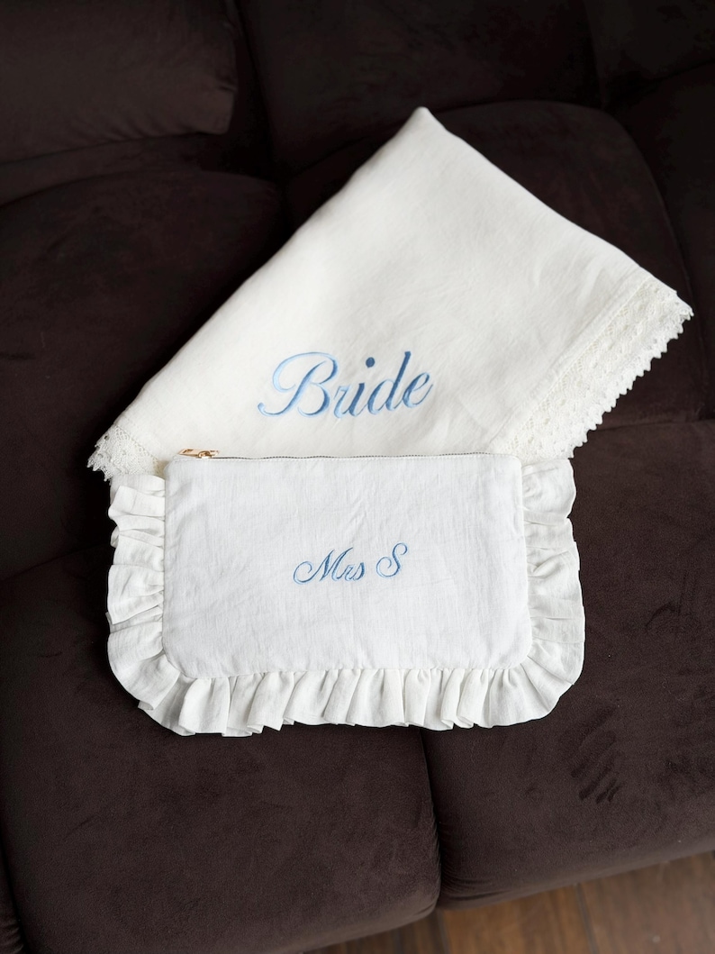 K&ouml;nnte beinhalten: Ein wei&szlig;es Braut-Set aus Leinen. Es besteht aus einem Kissenbezug mit dem blauen Schriftzug "Bride" und einer passenden Clutch mit der blauen Stickerei "Mrs S". Die Clutch hat einen R&uuml;schenrand.