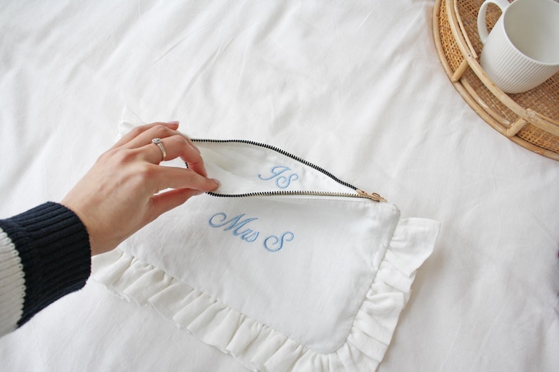 K&ouml;nnte beinhalten: Wei&szlig;e Leinen-Pochette mit R&uuml;schenkante und goldenem Rei&szlig;verschluss. Die Pochette ist mit den Initialen "IS" und "Mrs S" in hellblauer Farbe bestickt. Eine Hand &ouml;ffnet die Pochette.