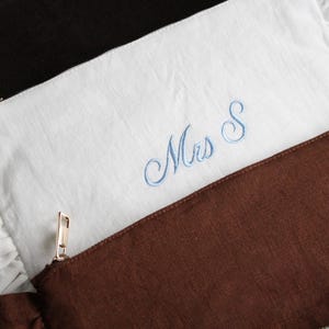 K&ouml;nnte beinhalten: Zwei Leinenbeutel, einer wei&szlig; und einer braun, sind &uuml;bereinander gelegt. Der wei&szlig;e Beutel hat eine hellblaue Stickerei "Mrs S" und einen ger&uuml;schten Rand. Beide Beutel haben goldene Rei&szlig;verschl&uuml;sse. Der braune Beutel ist unten sichtbar.
