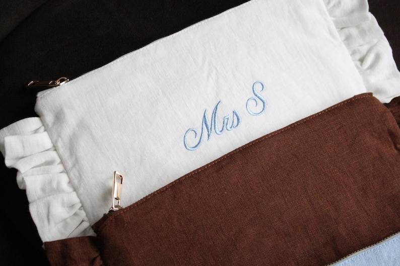 K&ouml;nnte beinhalten: Zwei Leinenbeutel, einer wei&szlig; und einer braun, sind &uuml;bereinander gelegt. Der wei&szlig;e Beutel hat eine hellblaue Stickerei "Mrs S" und einen ger&uuml;schten Rand. Beide Beutel haben goldene Rei&szlig;verschl&uuml;sse. Der braune Beutel ist unten sichtbar.