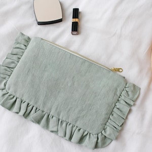 K&ouml;nnte beinhalten: Eine hellgr&uuml;ne rechteckige Clutch mit ger&uuml;schtem Rand und goldenem Rei&szlig;verschluss. Die Clutch liegt auf einer wei&szlig;en Oberfl&auml;che, neben einem Kompaktspiegel, Lippenstift und einem geflochtenen Tablett mit einer wei&szlig;en Tasse.