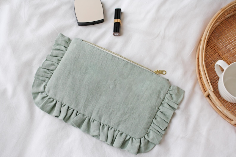 K&ouml;nnte beinhalten: Eine hellgr&uuml;ne rechteckige Clutch mit ger&uuml;schtem Rand und goldenem Rei&szlig;verschluss. Die Clutch liegt auf einer wei&szlig;en Oberfl&auml;che, neben einem Kompaktspiegel, Lippenstift und einem geflochtenen Tablett mit einer wei&szlig;en Tasse.
