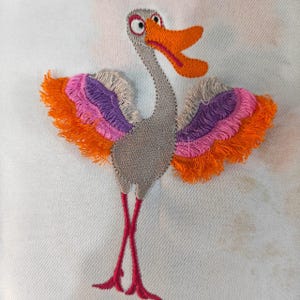 Op de afbeelding: Een geborduurde patch van een gestileerde vogel met een grijs lichaam, een feloranje snavel en lange dunne rode poten. De vleugels hebben gelaagde secties van oranje, roze en paarse draad met een gerafelde, getextureerde rand op witte stof.