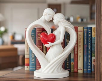 May include: A white ceramic sculpture of a couple embracing within a heart-shaped frame. The woman holds a red heart. The sculpture is on a wooden shelf with books in the background.