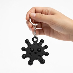 Pode incluir: Chaveiro preto com um design circular e espinhoso sorridente. O chaveiro é preso a uma corrente e um anel prateados. O design tem um rosto feliz e é feito de um material preto fosco. Um acessório divertido.