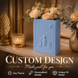 May include: A light blue rectangular vase featuring a relief design of a small figure, a fox, a rose under a glass dome, a crescent moon, and stars. The vase is filled with a bouquet of light peach roses and small white filler flowers.